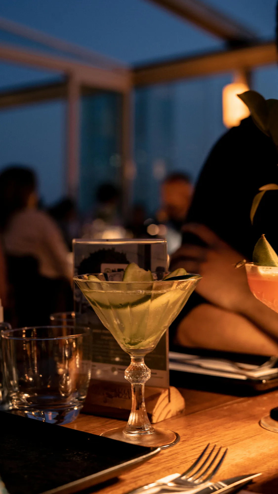 A cocktail on a rooftop bar counter at dusk, guests talking behind it