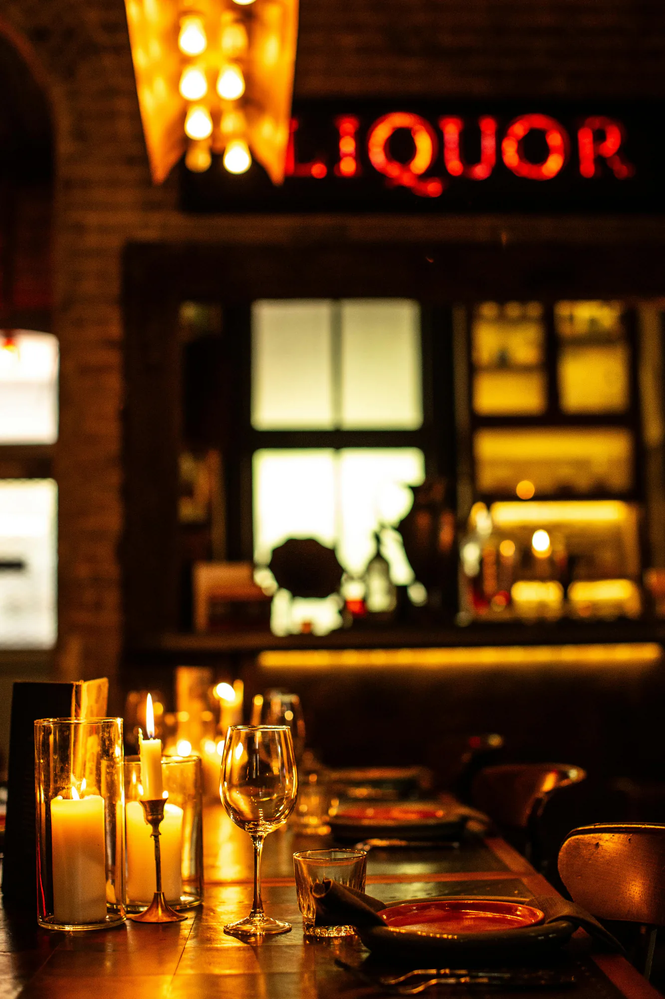 Candles and wine glasses on a bar under a glowing liquor sign