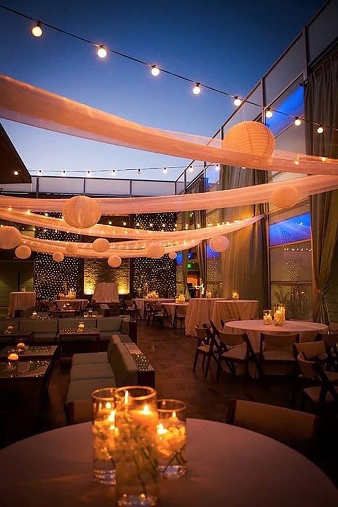 A rooftop lounge at blue hour with paper lanterns, draped fabric and candlelit tables