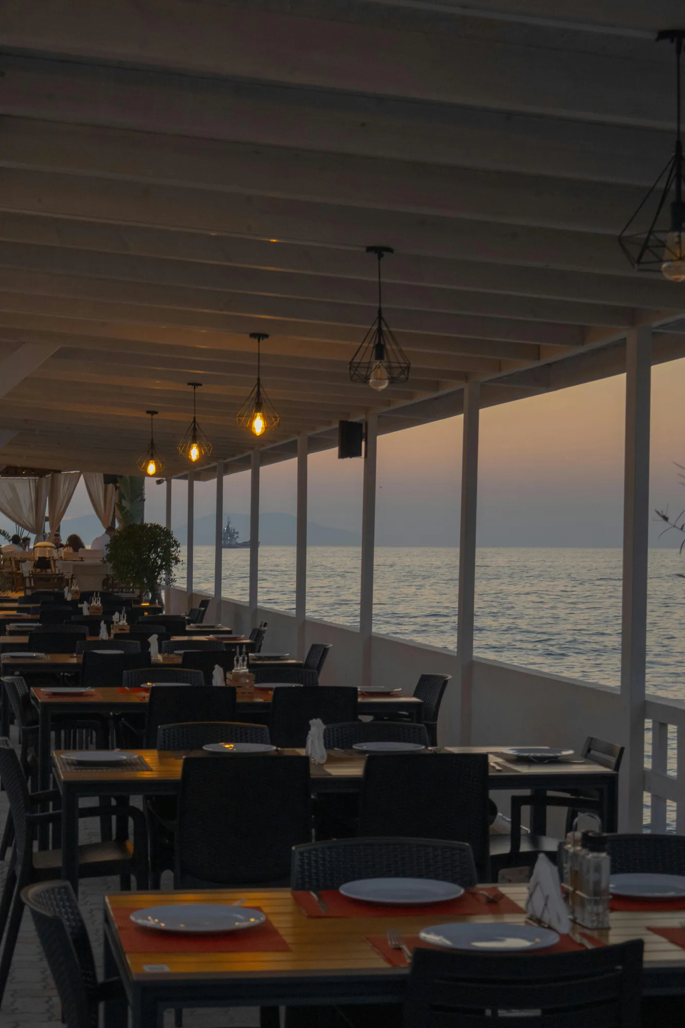 A covered seaside terrace with set tables and hanging lights at dusk